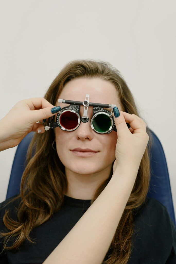 Unrecognizable oculist correcting phoropter with tinted lenses on eyes of female patient while doing vision check up against white wall